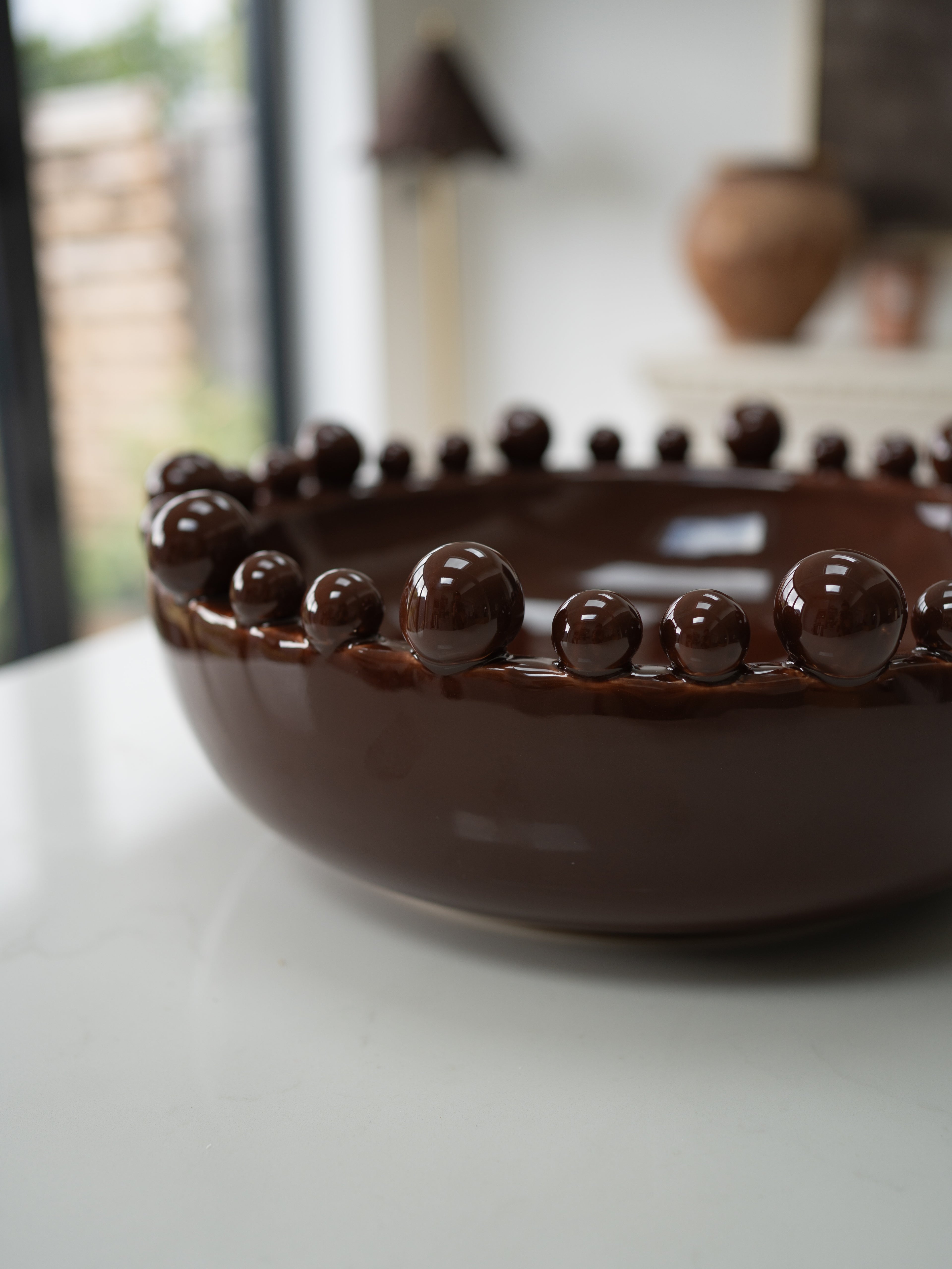A glossy dark brown ceramic bowl with a decorative border of raised spherical beads, photographed on a white tabletop with a softly blurred indoor background.
