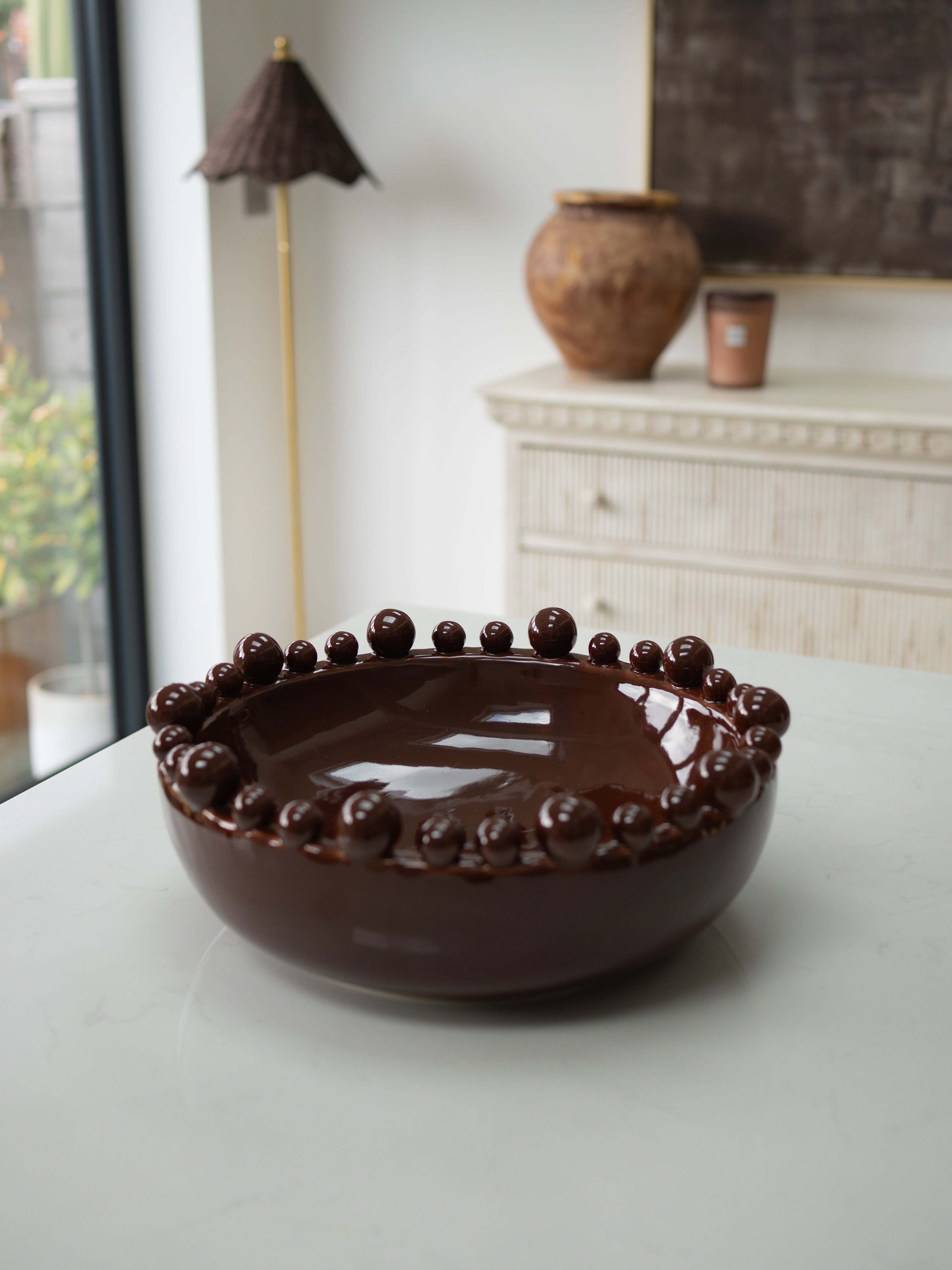 A dark brown ceramic bowl with a glossy finish and a decorative border of raised round beads, set on a light-colored table in a softly blurred interior room.