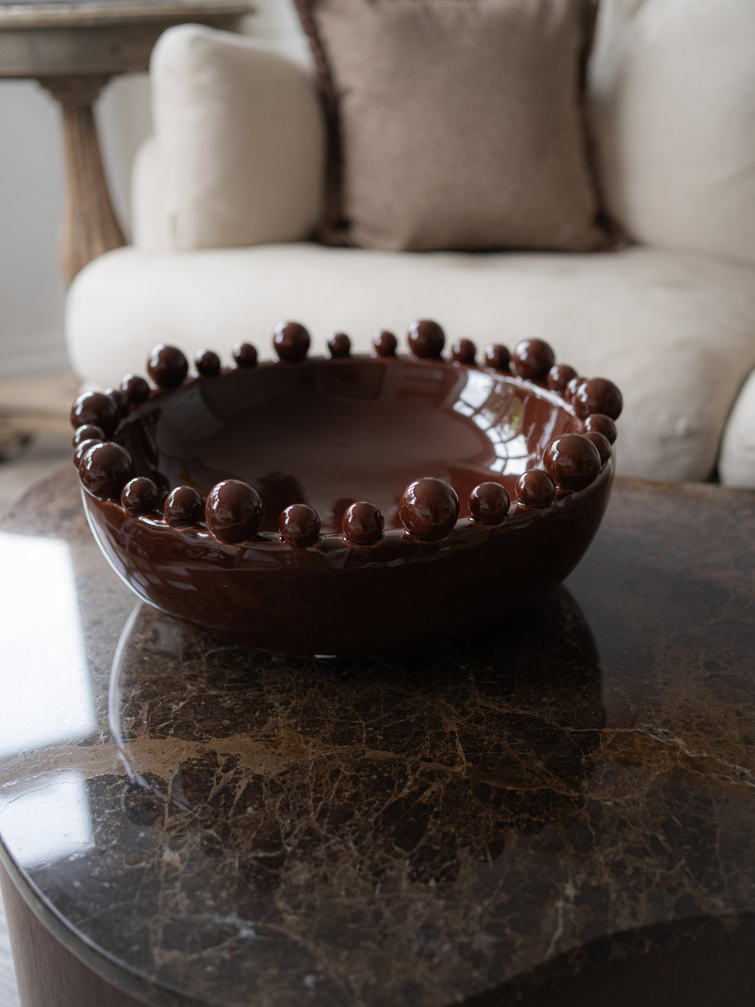 A dark brown glazed ceramic bowl with a raised beaded rim, displayed on a dark marble table in a living room setting with a sofa in the background.