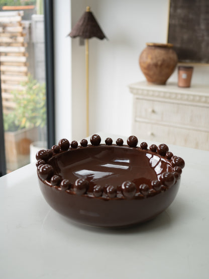 A glossy dark brown ceramic bowl with a raised beaded rim, displayed on a white tabletop in a softly lit interior setting.