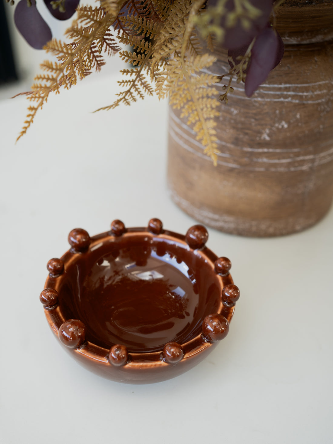 A small glossy brown ceramic bowl with a beaded rim, placed on a white surface with a rustic brown vase and dried foliage in the background.