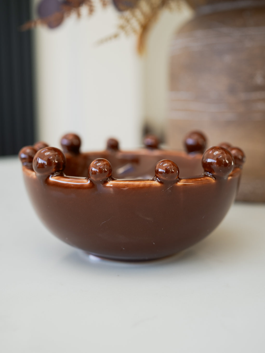 A glossy brown ceramic bowl with a scalloped rim and raised decorative beads, shown on a light tabletop with a softly blurred background.