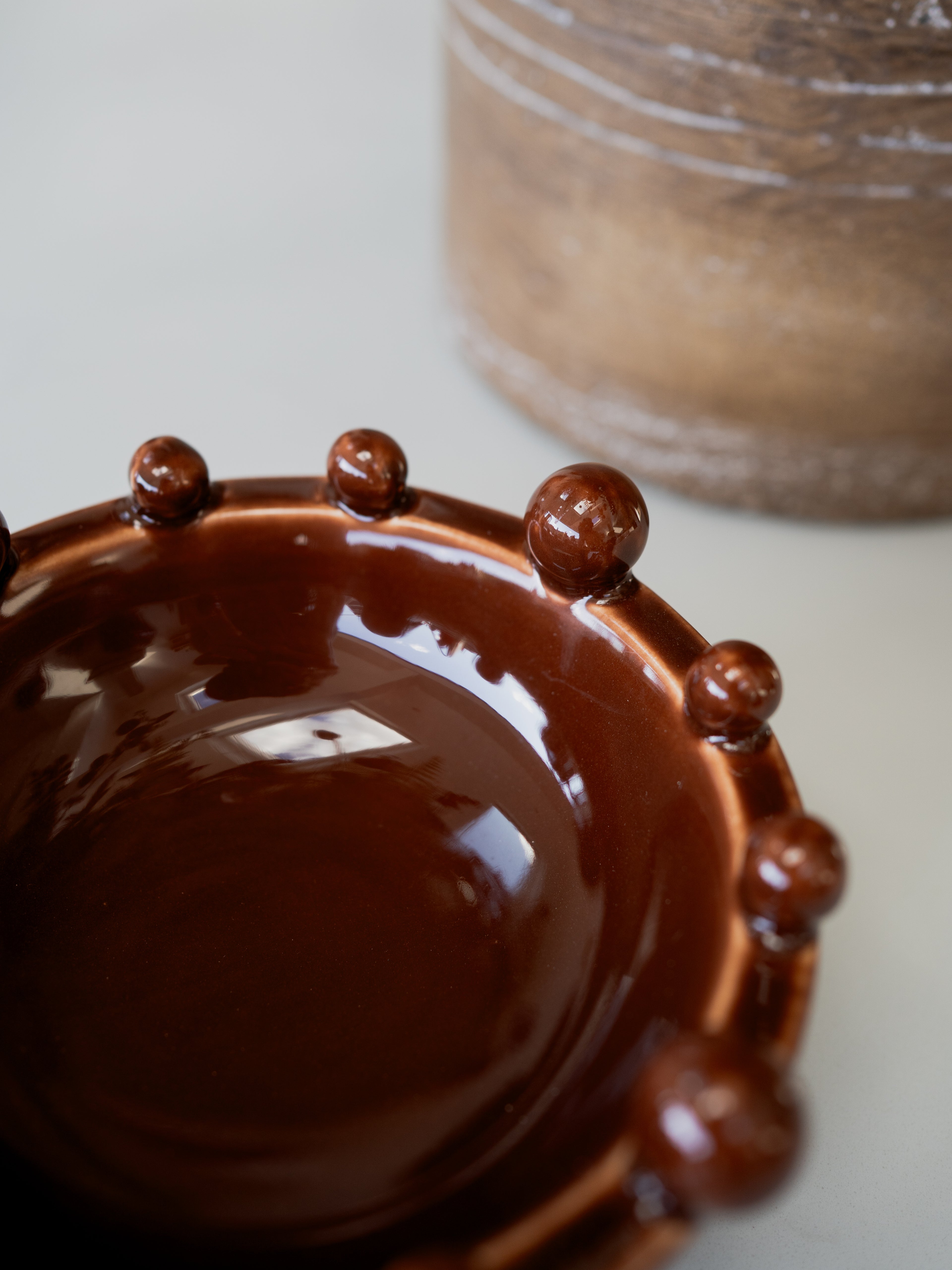 A close-up of a glossy dark brown ceramic bowl with a raised beaded rim, shown on a light neutral surface with a blurred brown vessel in the background.