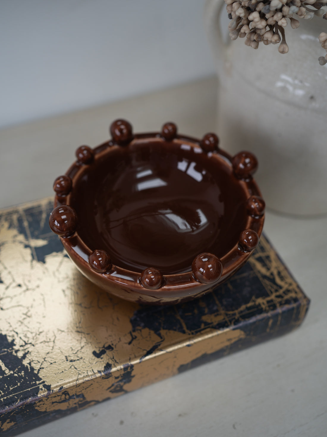 A small glossy brown ceramic bowl with a raised beaded rim, displayed on a distressed gold and navy box with a white vase in the background.