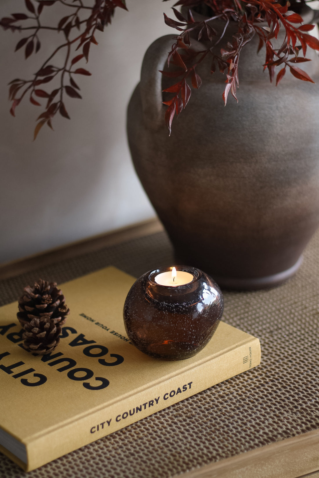 Candle in a glass holder on a book with a vase and branches in the background