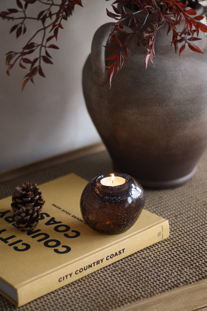 Candle in a glass holder on a book with a vase and branches in the background