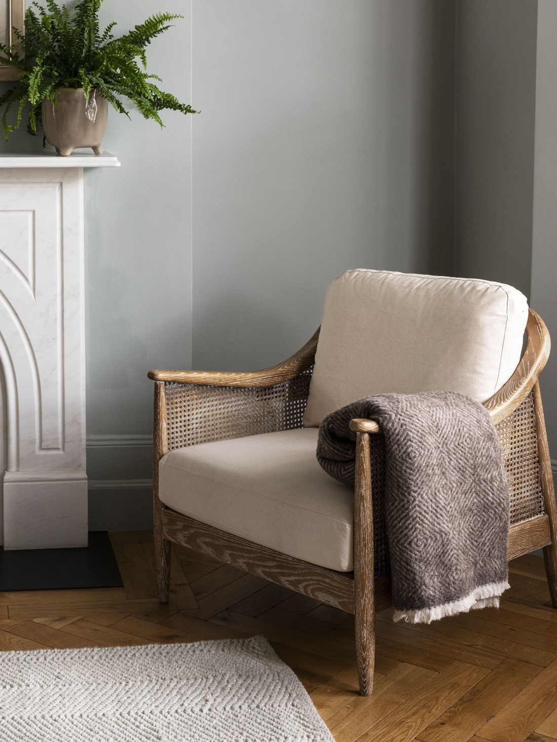 Wicker armchair with beige cushion and gray throw blanket in a room with a white fireplace and green plant.
