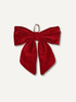 A large red velvet bow christmas decoration with a small metal loop at the top, displayed against a light off-white background.