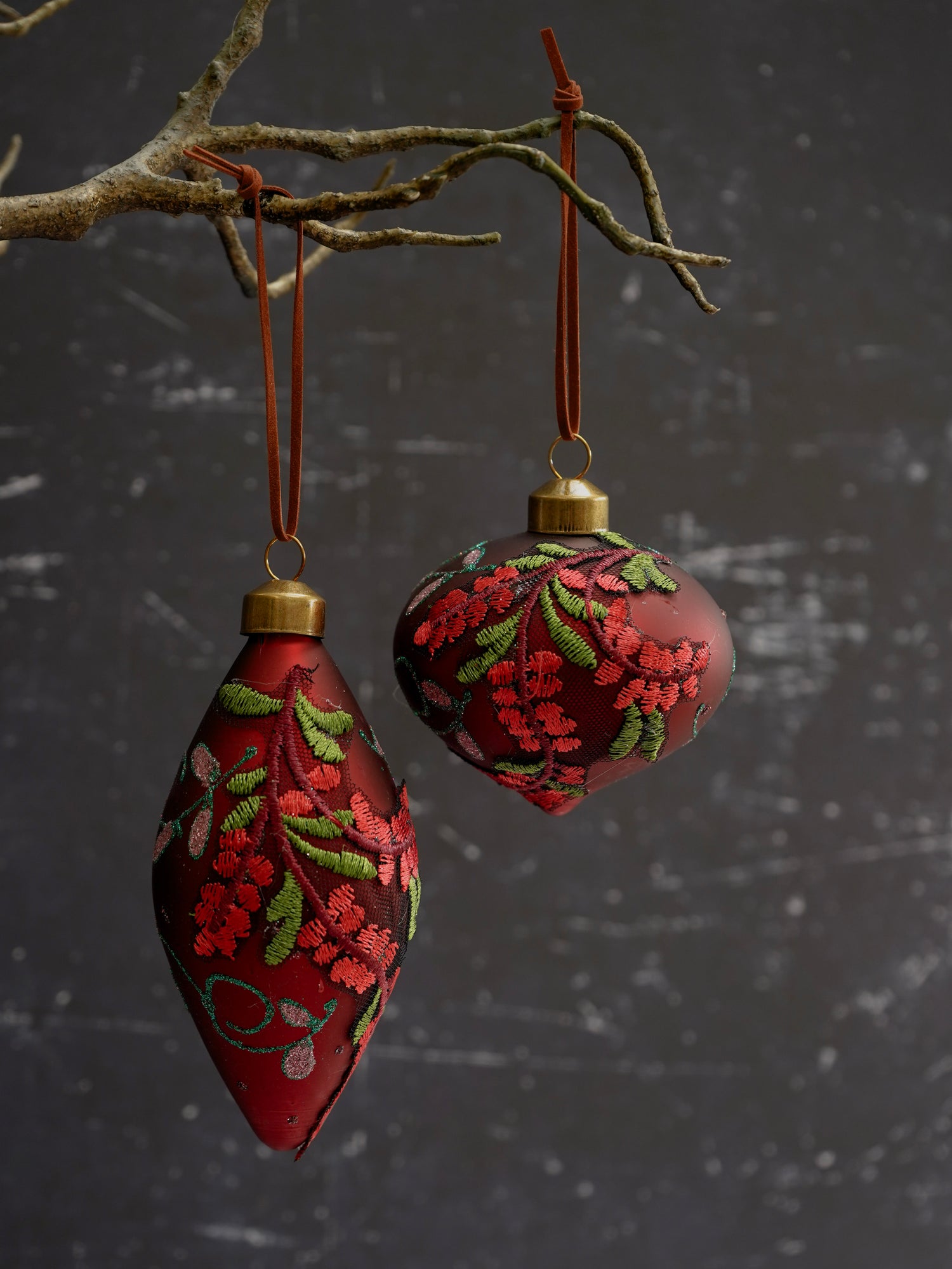Two red Christmas ornaments with floral patterns hanging from a branch against a dark textured background.