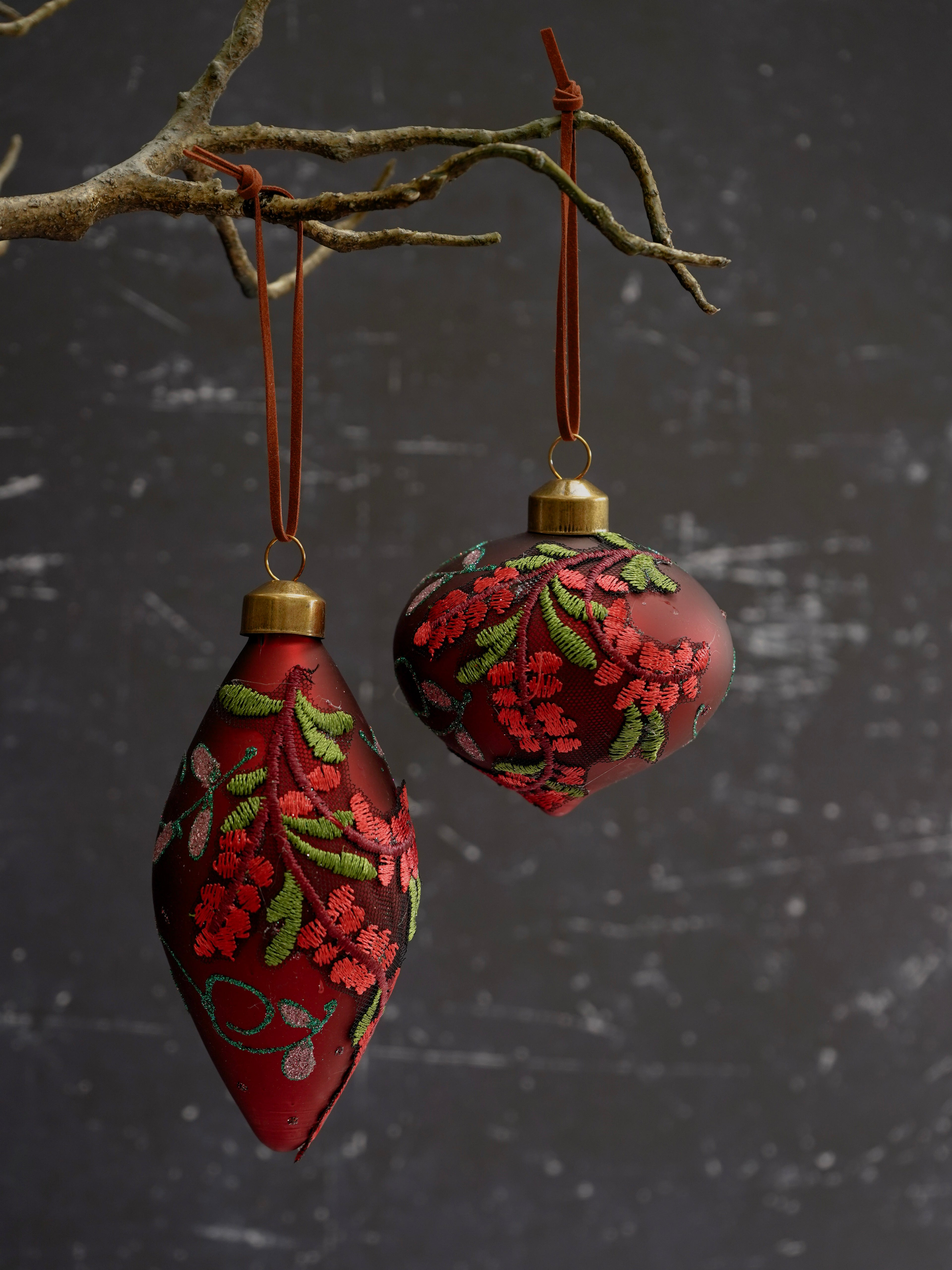 Two red Christmas ornaments with floral patterns hanging from a branch against a dark textured background.