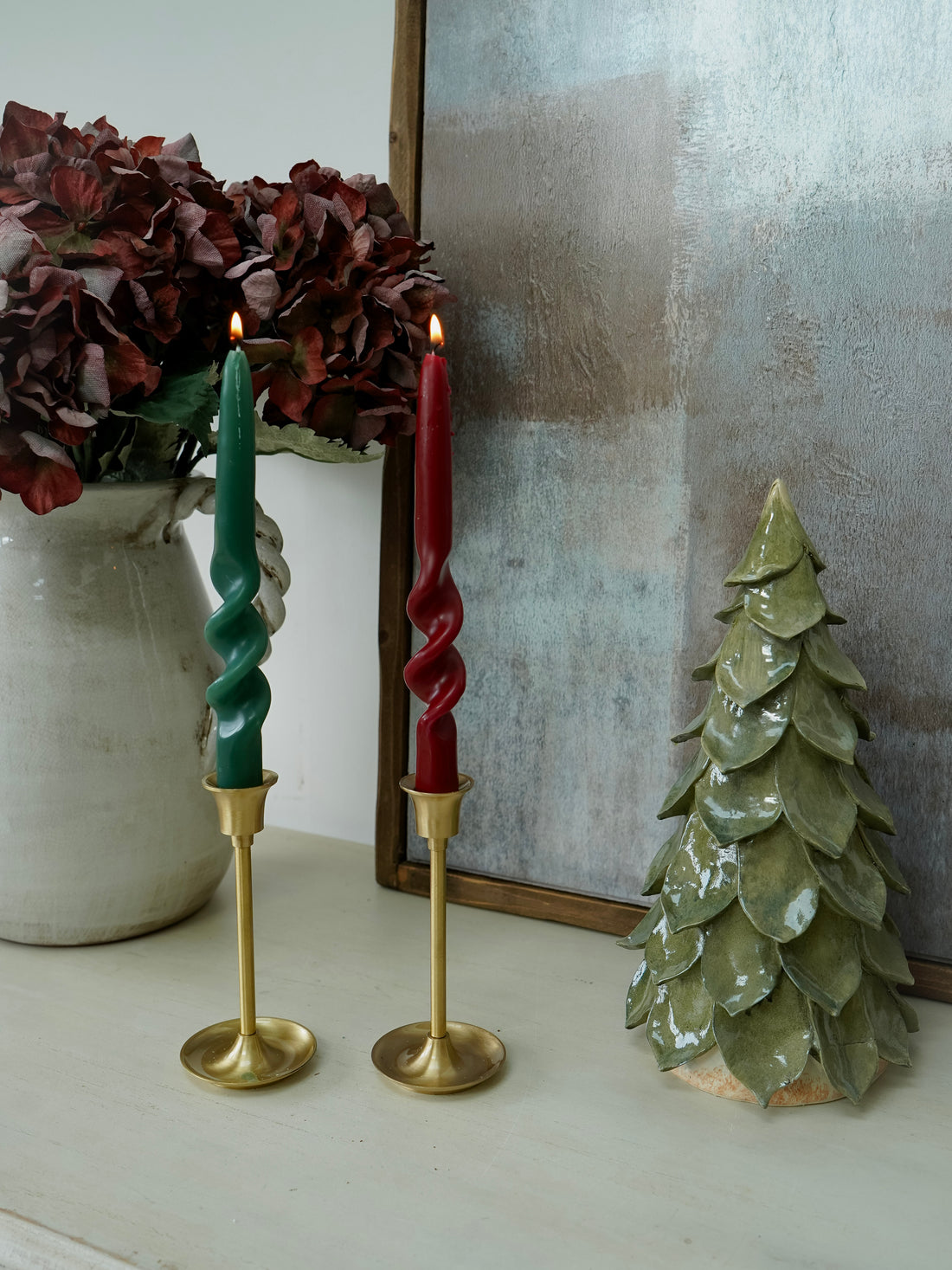Two twisted candles in gold holders with a decorative tree and flowers on a white surface.