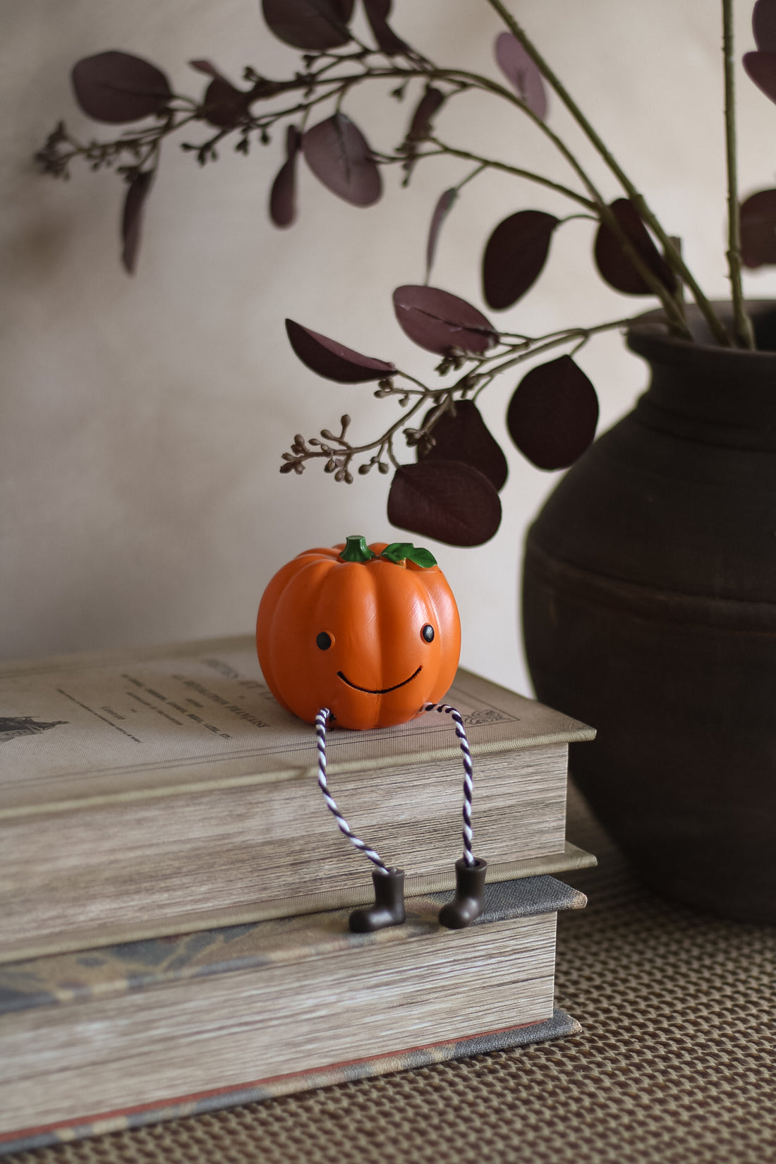 Small pumpkin figurine with a face and legs on top of books, with a vase and branches in the background.