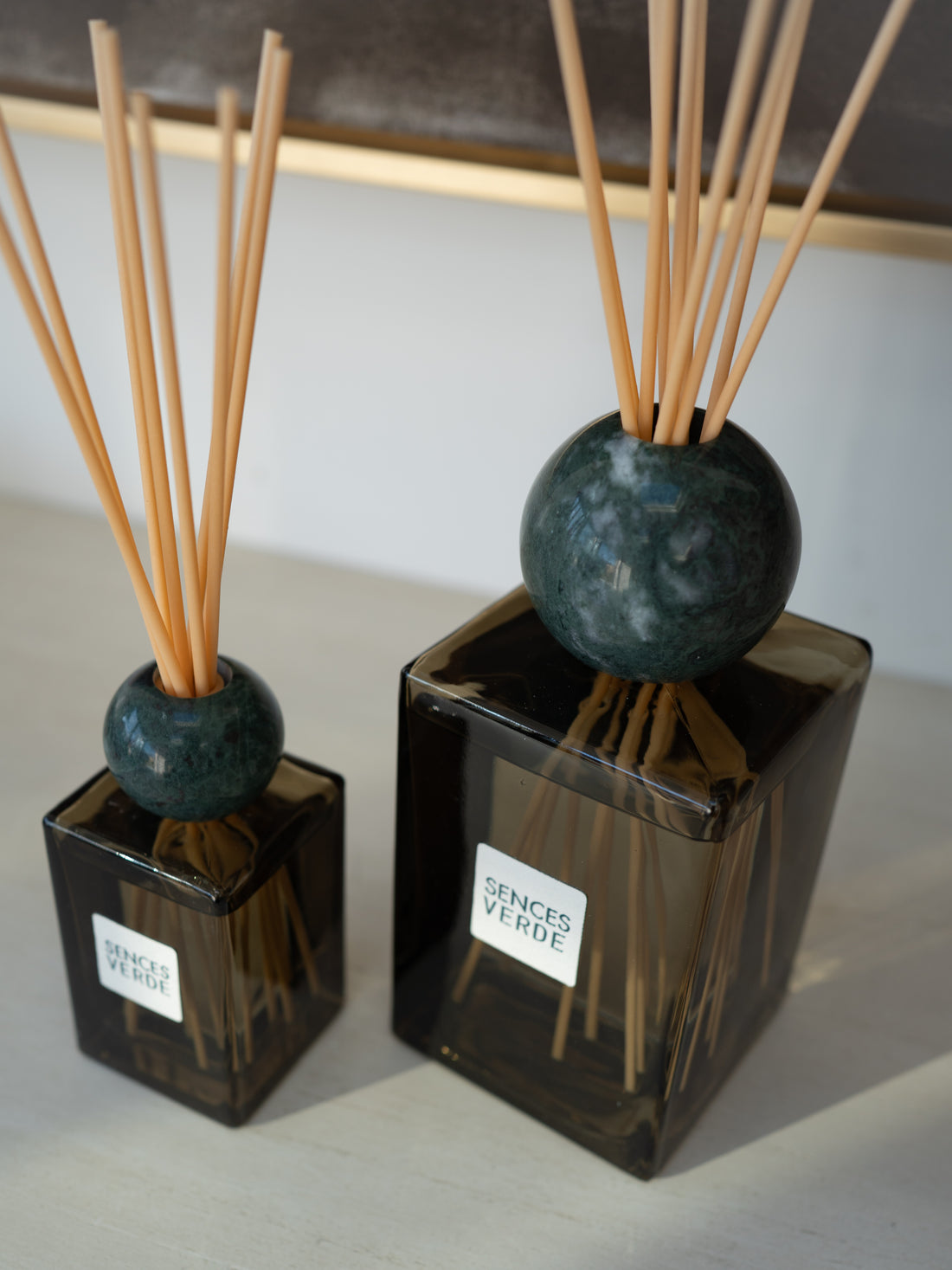 Two reed diffusers with dark green glass bases, round dark teal stone spheres, and natural wood reeds, with one larger diffuser in the foreground and a visible label reading &