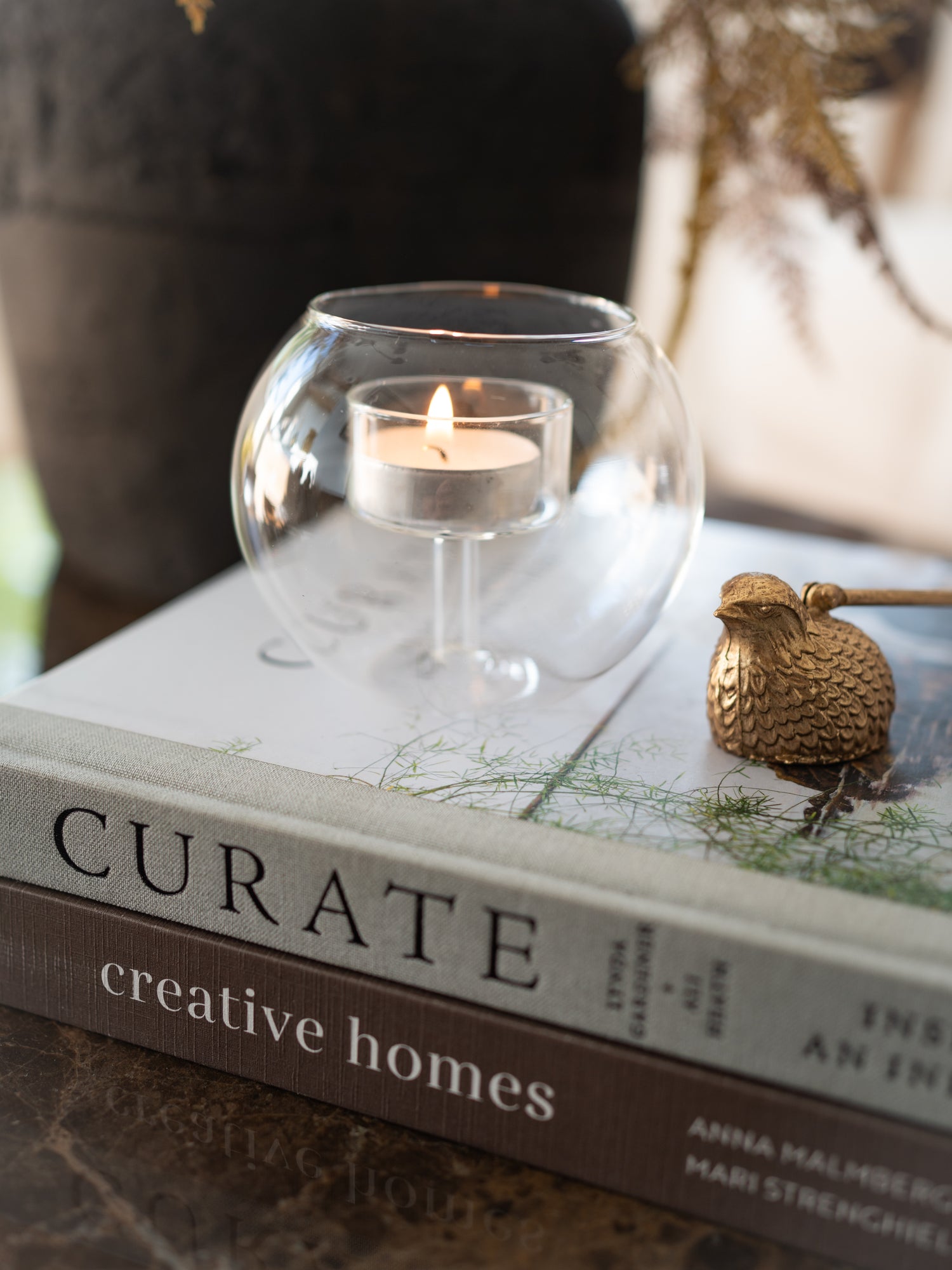 A clear glass candle holder containing a lit tea light candle, styled on top of a book with a small gold decorative bird candle snuffer beside it.