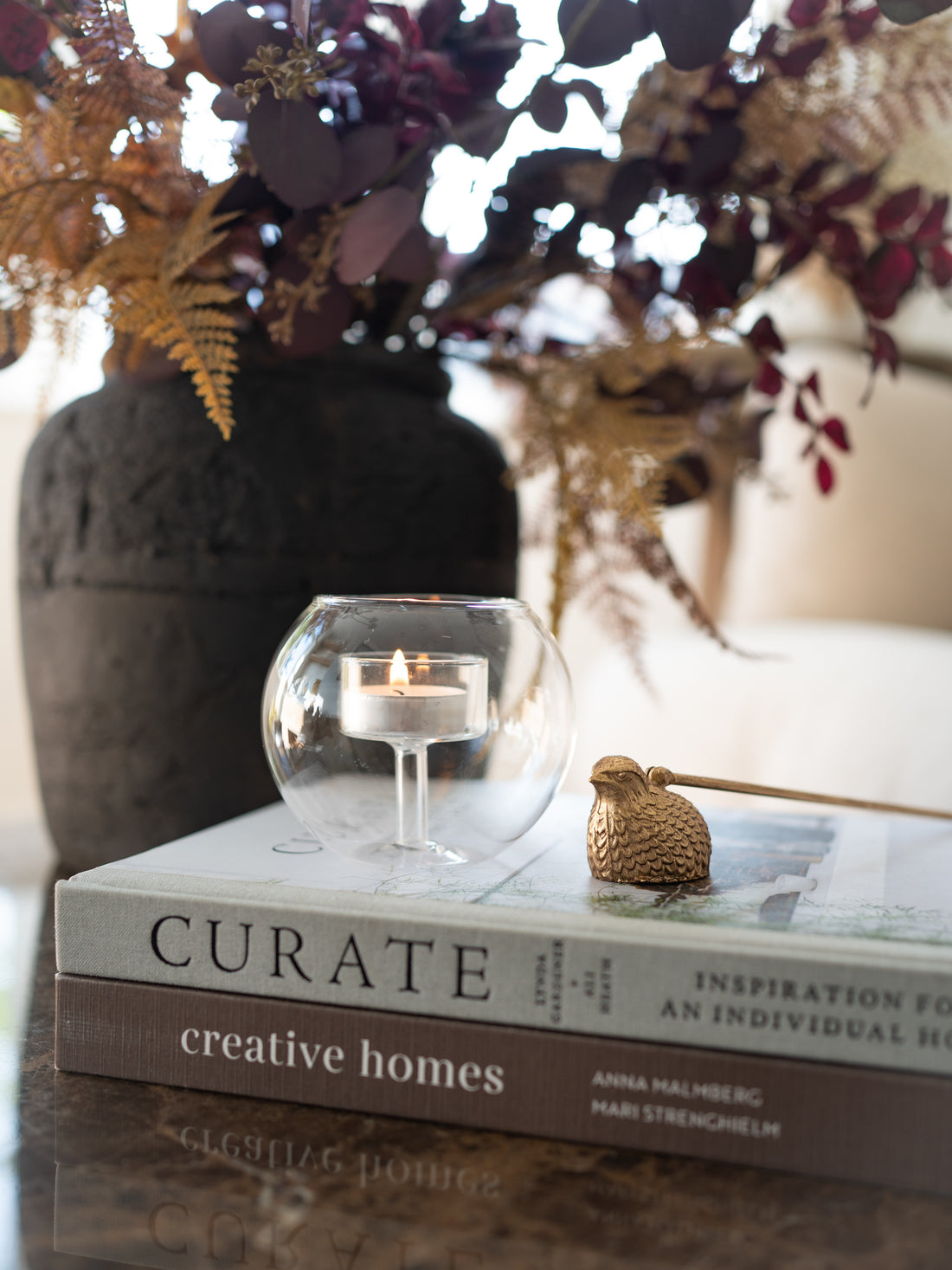 A lit tealight candle in a clear glass holder resting on a stack of books, with a small gold bird figurine nearby and a dark vase of dried flowers in the background.