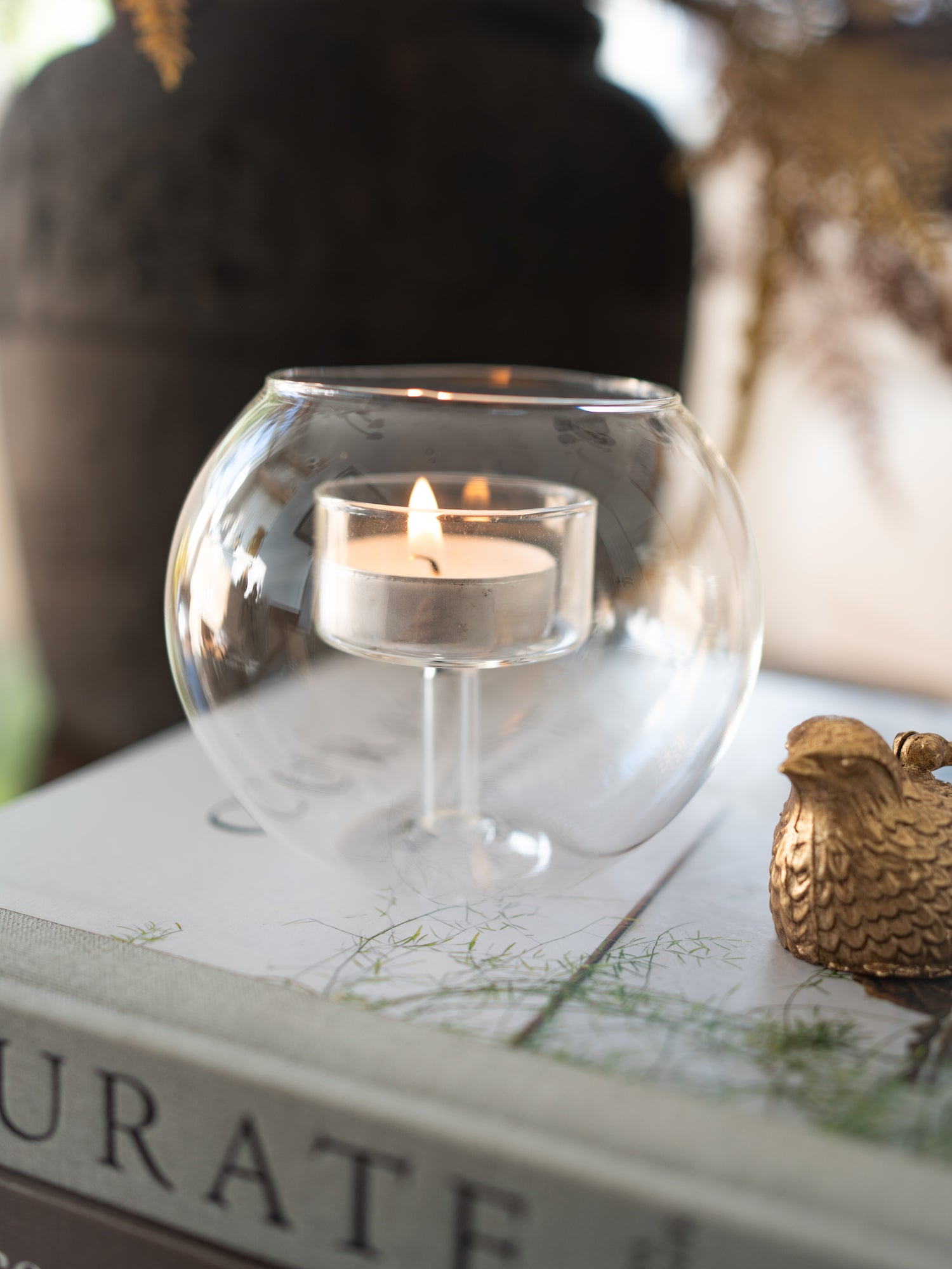 A clear glass fishbowl-shaped candle holder with a lit tealight candle inside, styled on top of a book with a small gold decorative bird figurine nearby.