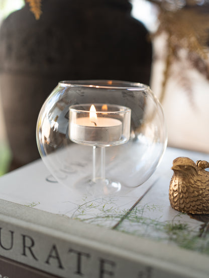A clear glass fishbowl-shaped candle holder with a lit tealight candle inside, styled on top of a book with a small gold decorative bird figurine nearby.