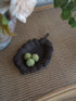 A dark brown leaf-shaped dish holding three small green spiky decorative balls, placed on a woven tan mat with blurred flowers and greenery in the background.