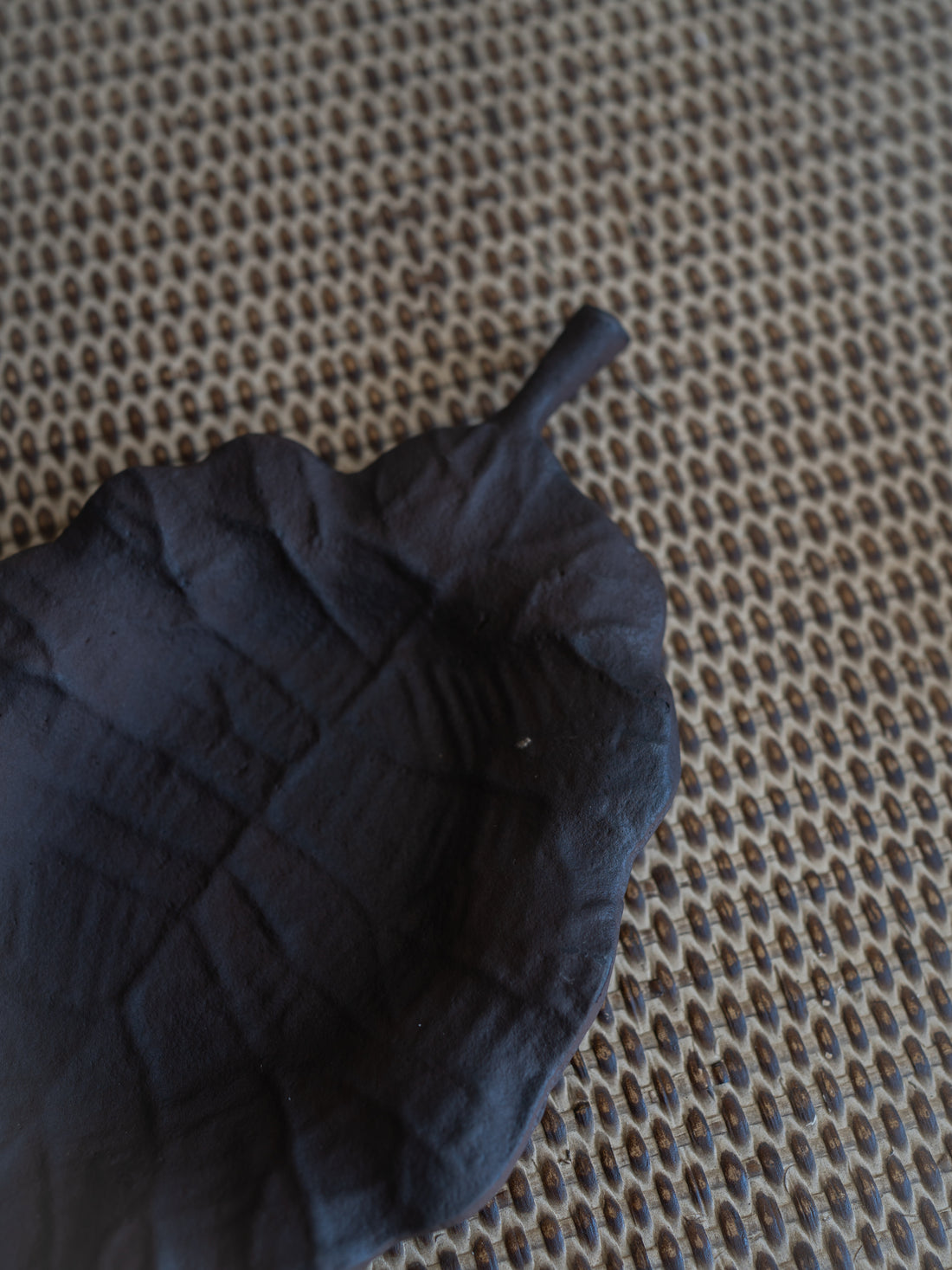 A dark, matte leaf-shaped object with a textured surface and short stem, placed on a woven beige and brown textured background.