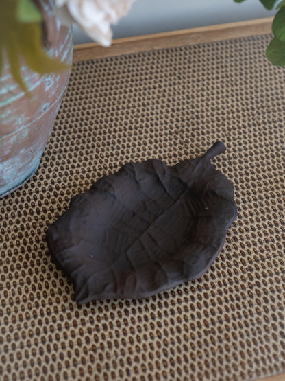 A dark brown leaf-shaped dish or tray with a textured, embossed leaf pattern, resting on a woven brown mat.