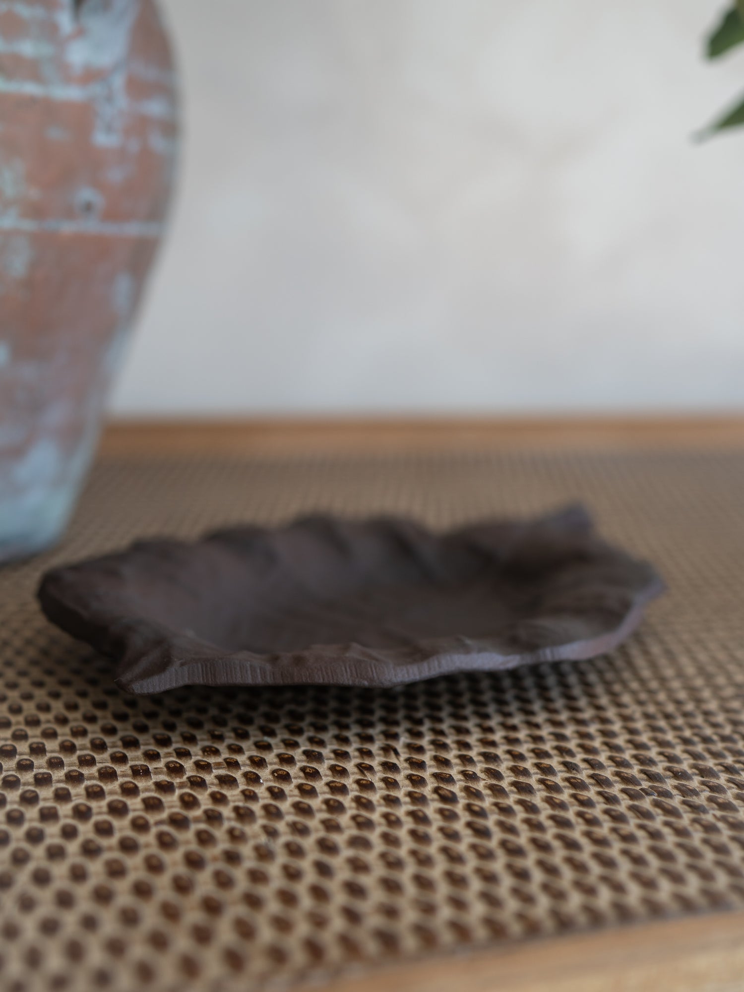 A small, rustic dark brown metal dish with a wavy, leaf-like rim, resting on a woven brown mat with a blurred vase in the background.