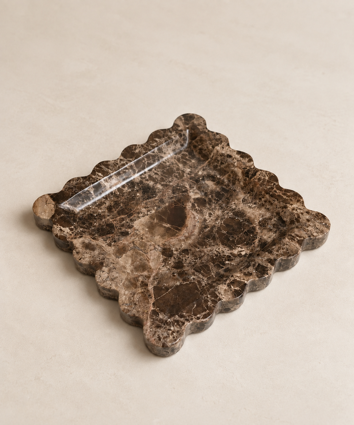 A square dark brown marble tray with a scalloped edge and a clear glass insert, photographed on a light beige textured background.
