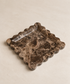 A square dark brown marble tray with a scalloped edge and a clear glass insert, photographed on a light beige textured background.
