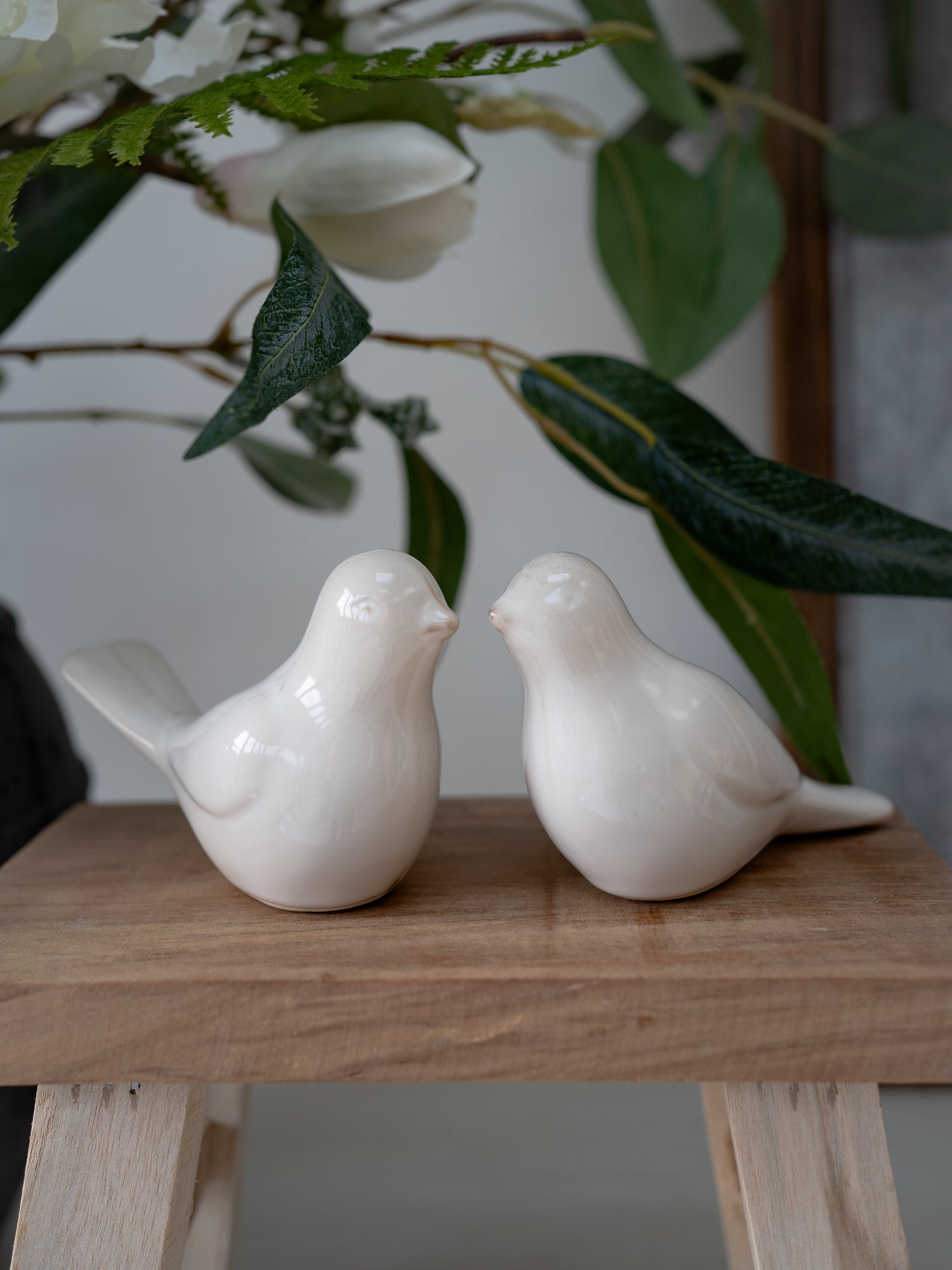 Two white ceramic birds on a wooden surface with green leaves in the background