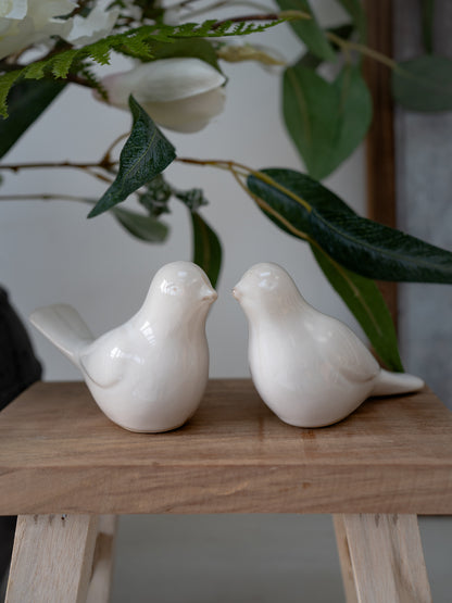 Two white ceramic birds on a wooden surface with green leaves in the background