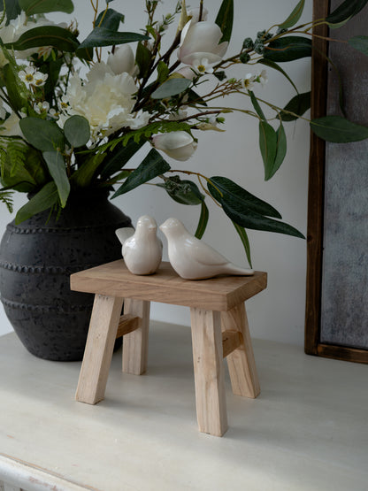 Decorative scene with a vase of flowers, a wooden stool, and two white birds on a neutral background.
