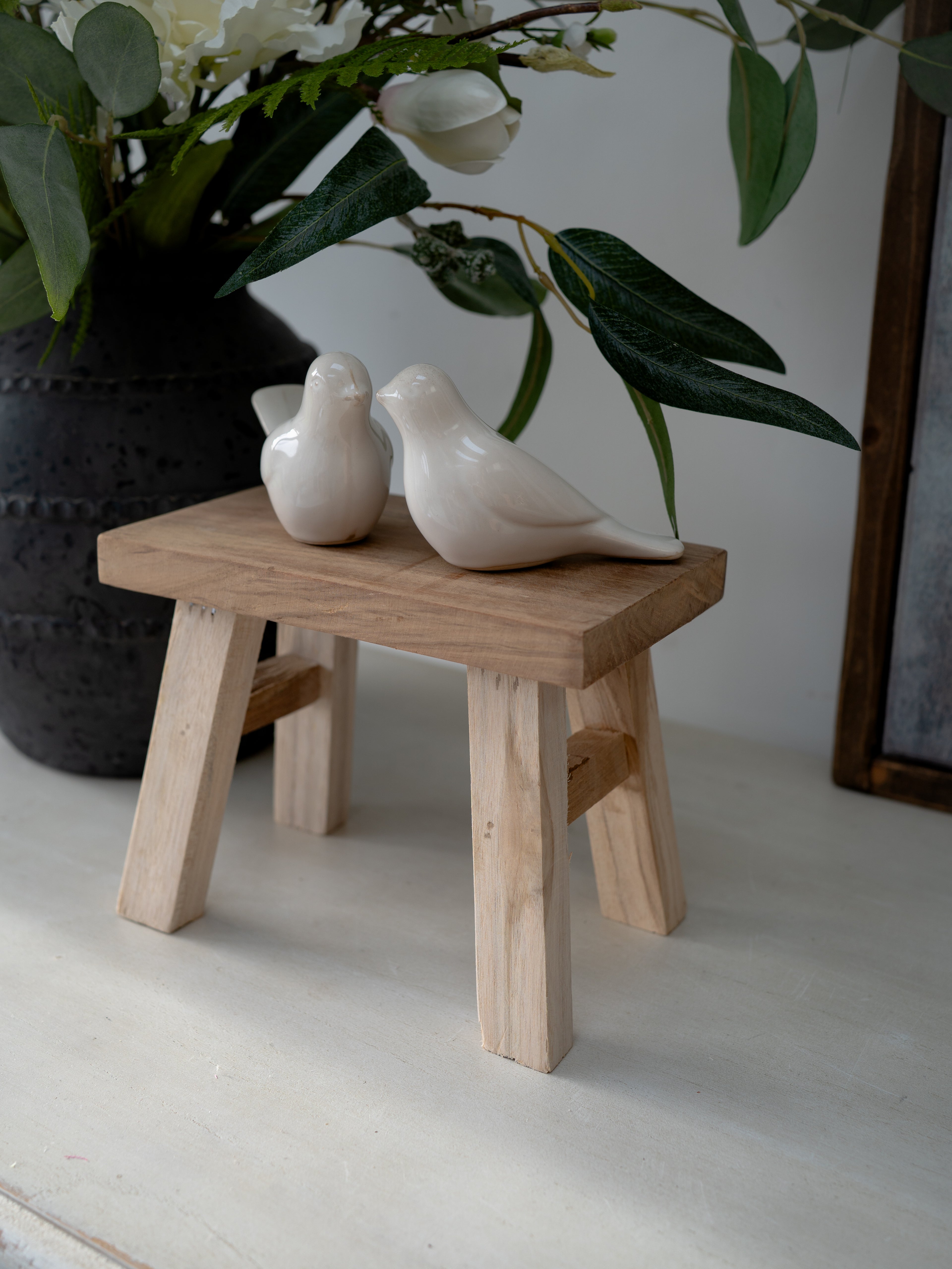 Two ceramic birds on a small wooden stool with a vase of flowers in the background.