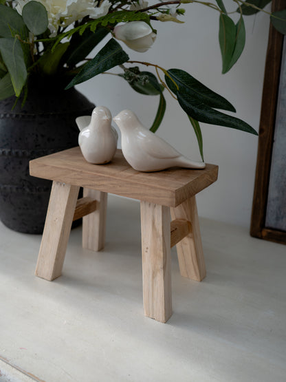Two ceramic birds on a small wooden stool with a vase of flowers in the background.