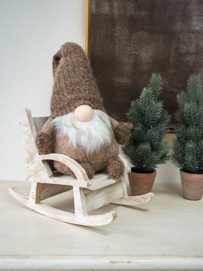 A brown knit gonk doll with a white beard and pink nose sits on a small white rocking chair. Two small potted evergreen trees are behind it against a neutral indoor background.