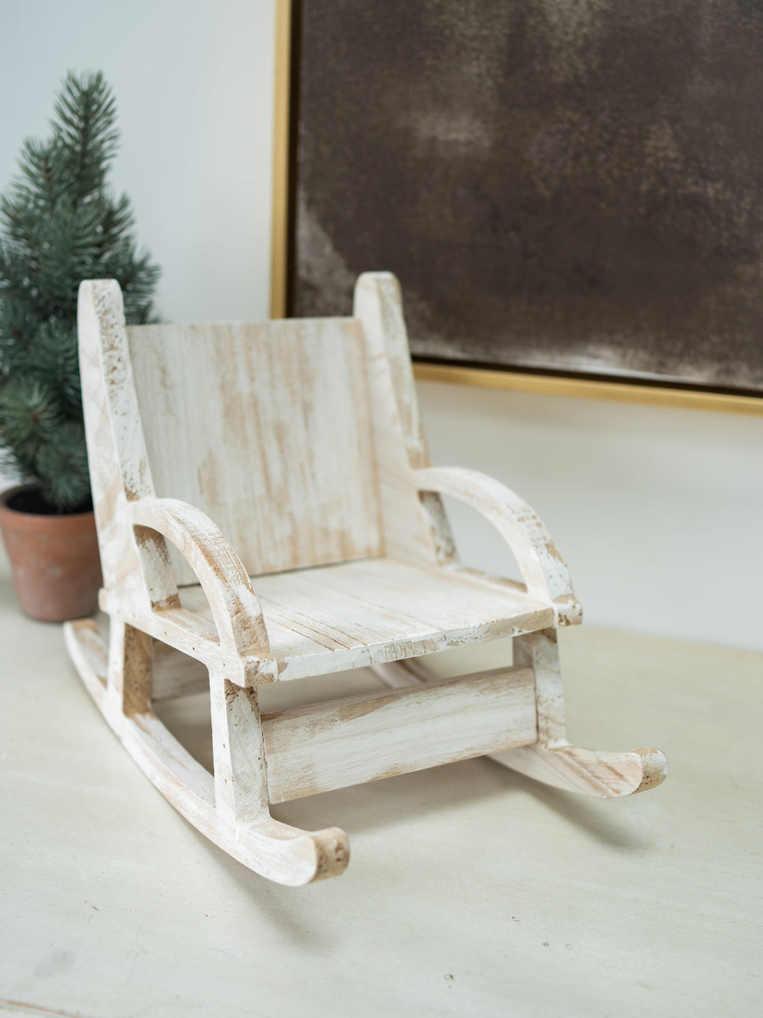 A miniature distressed white wooden rocking chair with a rustic, weathered finish, styled on a tabletop next to a small potted pine tree.