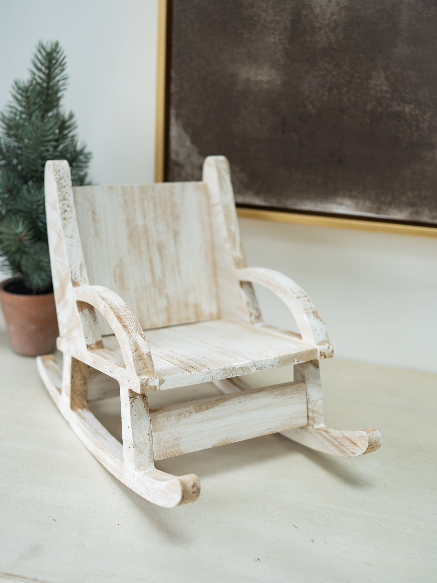 A miniature distressed white wooden rocking chair with a rustic, weathered finish, styled on a tabletop next to a small potted pine tree.