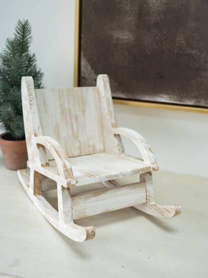 A miniature distressed white wooden rocking chair with a rustic, weathered finish, styled on a tabletop next to a small potted pine tree.