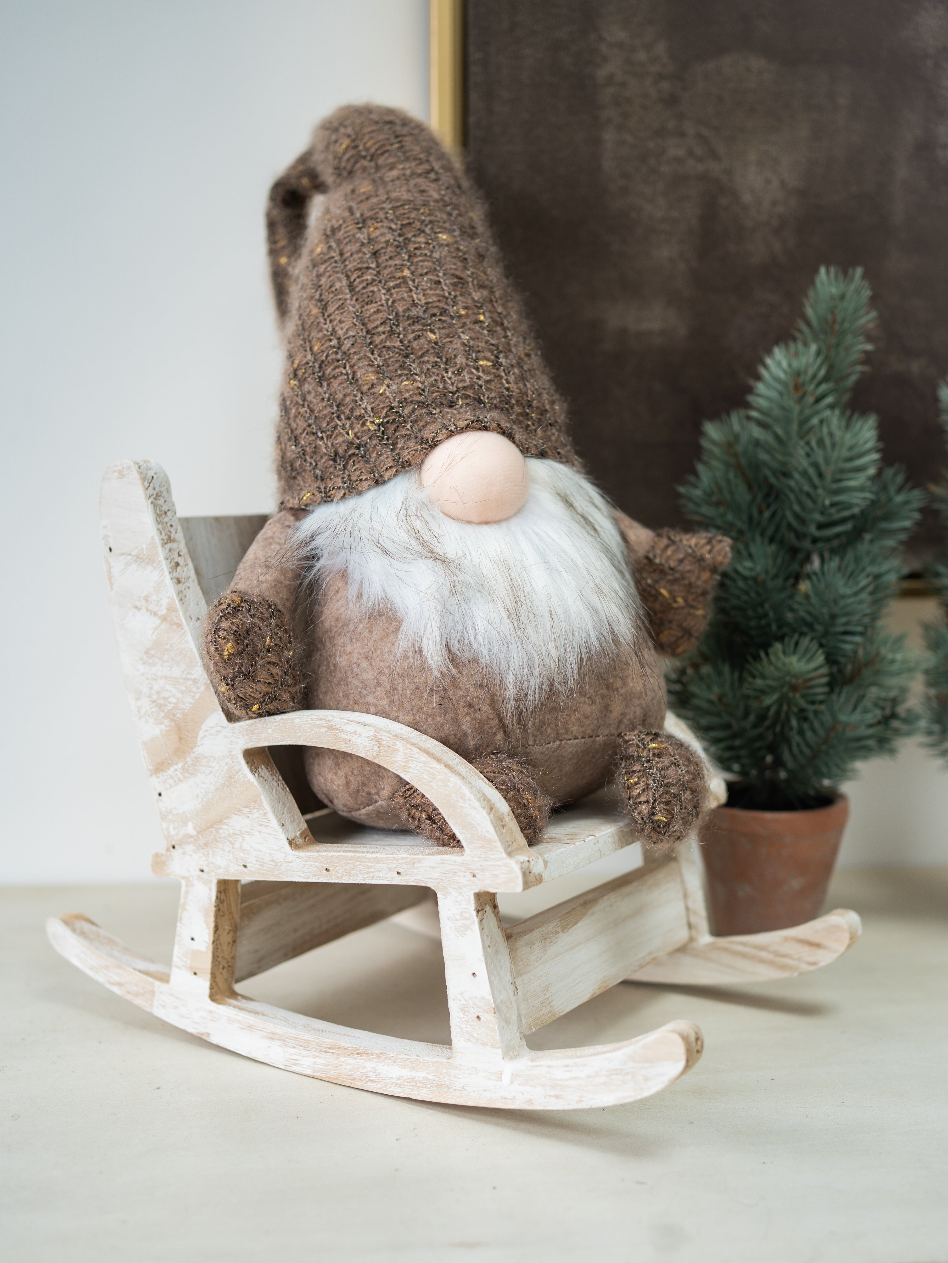 A rustic brown knit gonk doll with a white faux-fur beard and pink nose, seated in a distressed white wooden rocking chair. A small potted evergreen tree stands in the background against a neutral indoor setting.