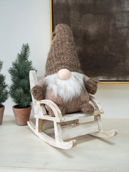 A plush Christmas gonk doll with a brown knit hat, white beard, and pink nose sits on a small white wooden rocking chair. The scene is styled with potted evergreen trees against a neutral wall.