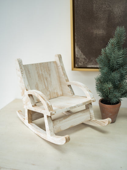 A miniature distressed white wooden rocking chair toy, styled next to a small potted green pine tree on a light beige surface against a white wall.