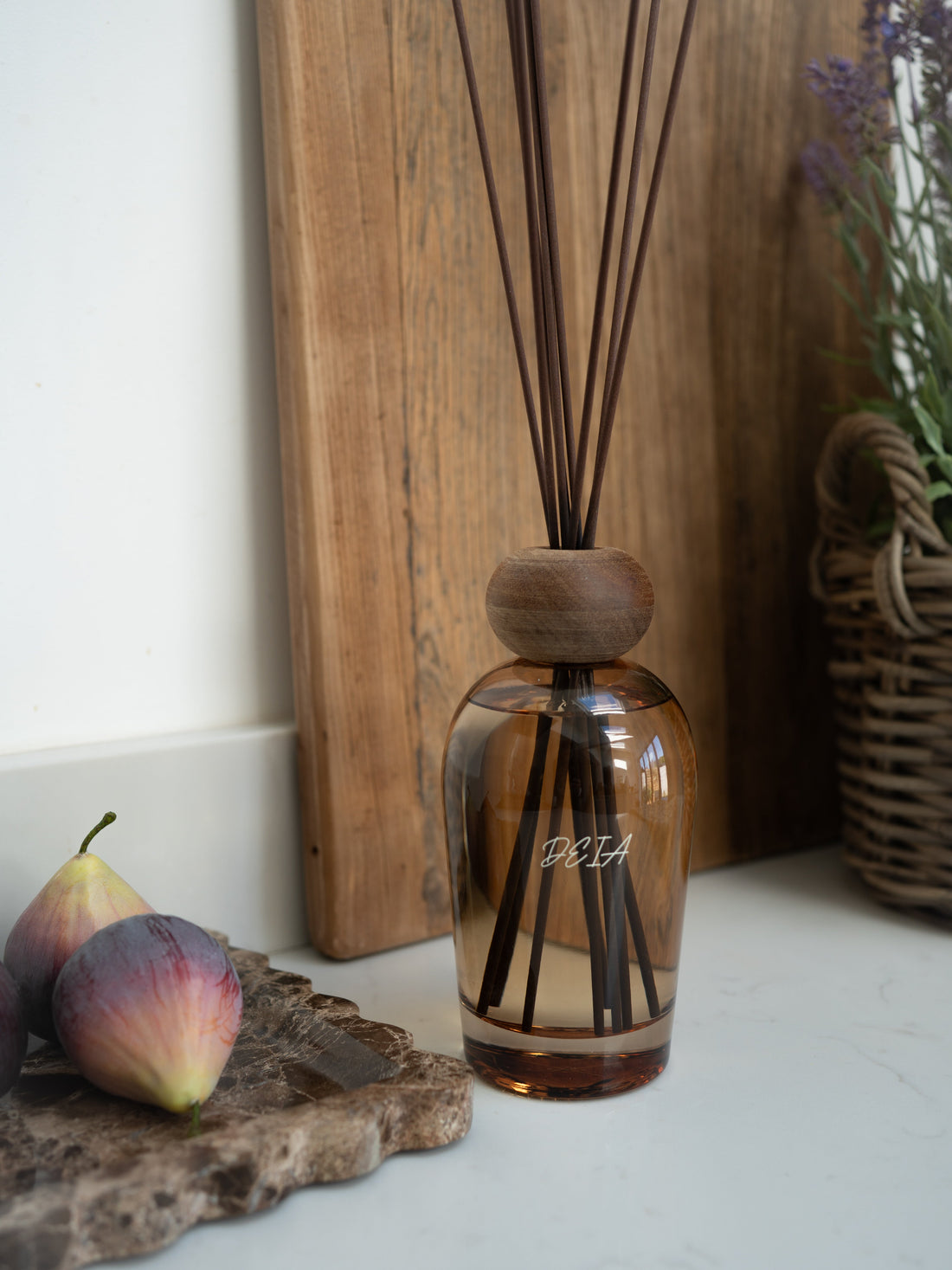 A reed diffuser in brown glass bottle with brown stick and wooden cap and Deia on a surface with fruits and a basket in the background.