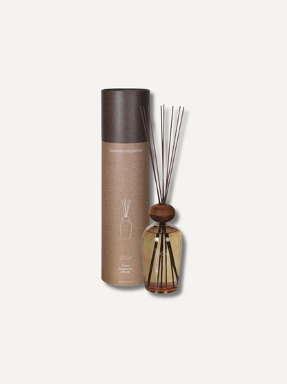 A statement reed diffuser set with a cylindrical kraft paper bottle, dark brown cap, amber liquid, and wooden reeds, shown next to its matching cardboard packaging on a light off-white background.