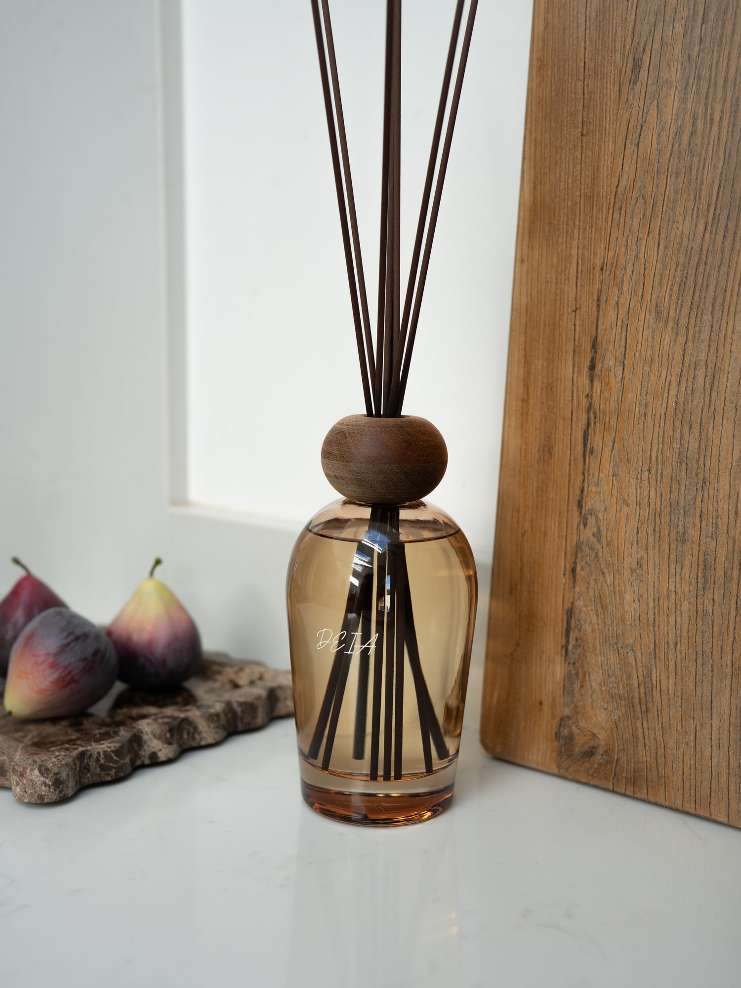 Brown diffuser bottle with wooden cap and sticks on a white surface with a wooden board and fruits in the background.