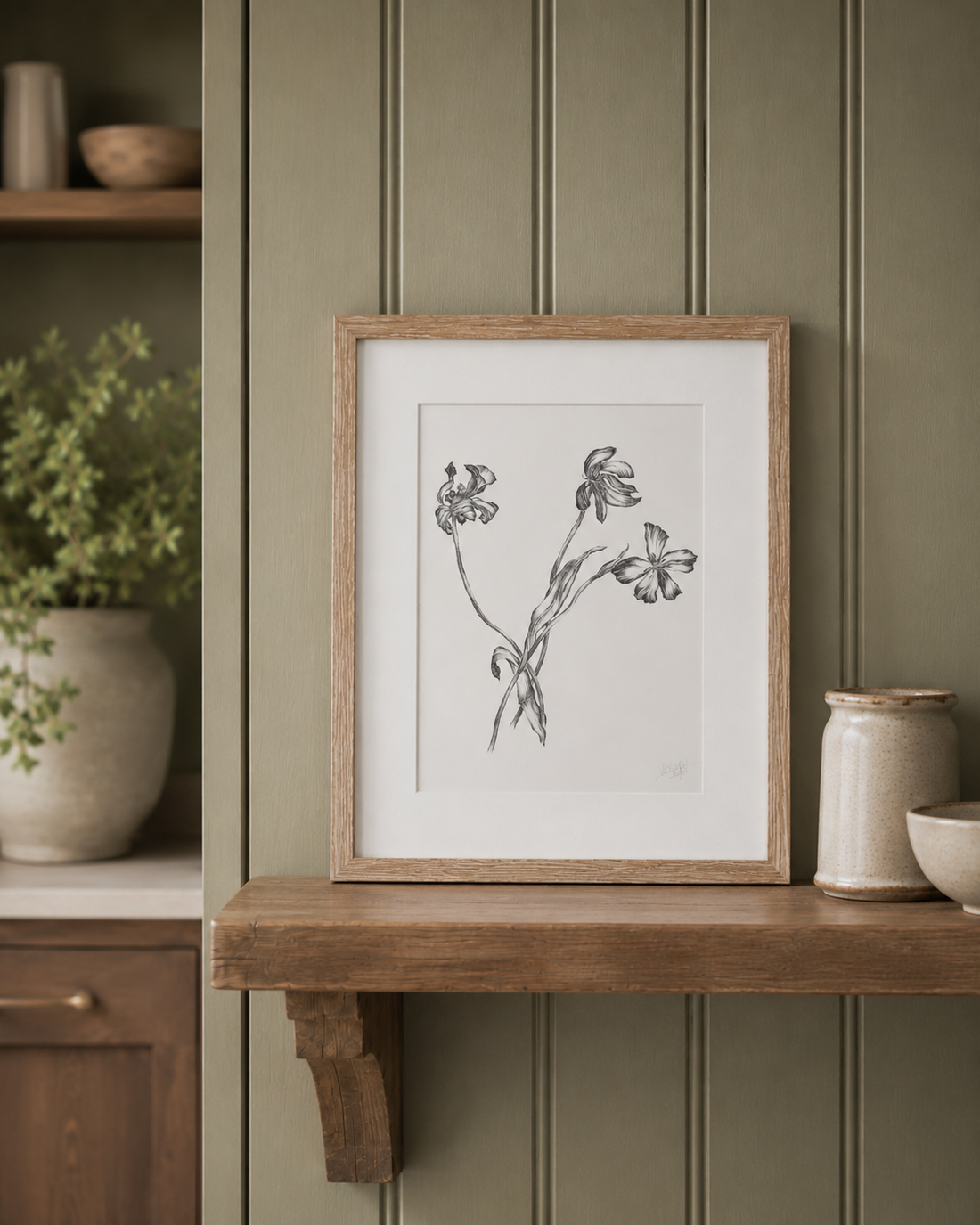 A framed botanical art print featuring black ink sketches of wildflowers on a white background, styled on a wooden shelf against a green paneled wall.