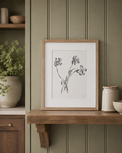 A framed botanical art print featuring black ink sketches of wildflowers on a white background, styled on a wooden shelf against a green paneled wall.
