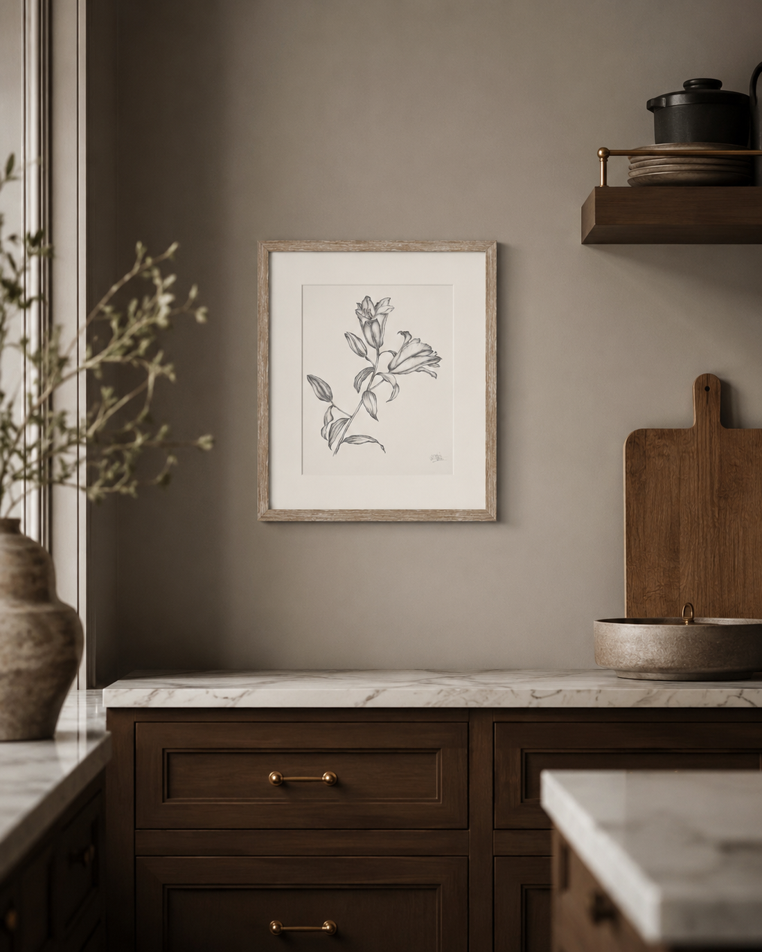 A framed botanical line drawing of lilies in a light wood frame, displayed on a warm taupe-gray wall in a styled kitchen setting with dark wood cabinets and marble countertops.