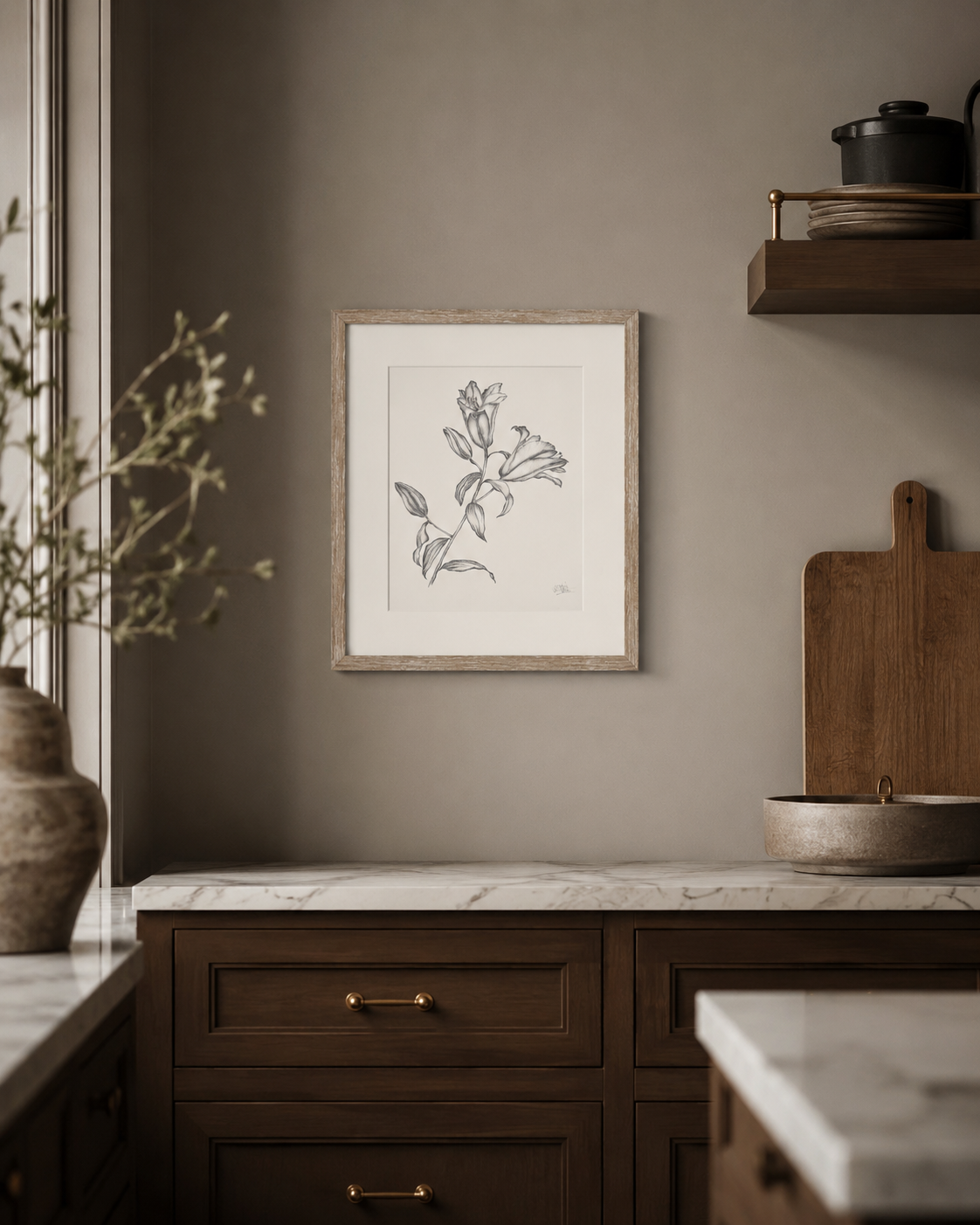A framed botanical line drawing of lilies in a light wood frame, displayed on a warm taupe-gray wall in a styled kitchen setting with dark wood cabinets and marble countertops.