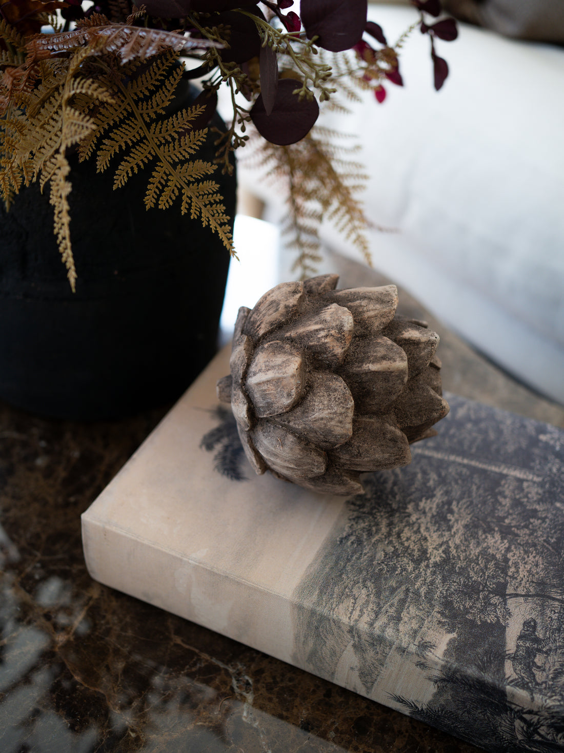 A decorative rustic pinecone-style decor piece resting on a book on a marble surface, with a black vase of dried ferns and foliage in the background.