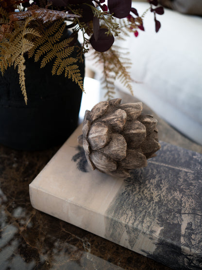 A decorative rustic pinecone-style decor piece resting on a book on a marble surface, with a black vase of dried ferns and foliage in the background.