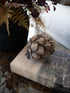 A decorative rustic pinecone-style decor piece resting on a book on a marble surface, with a black vase of dried ferns and foliage in the background.