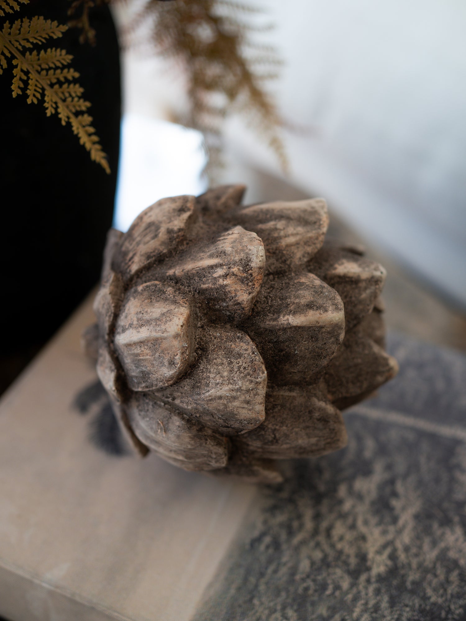A decorative faux pinecone sculpture in a weathered brown and gray stone-like finish, styled on a tabletop with blurred ferns and a neutral background.